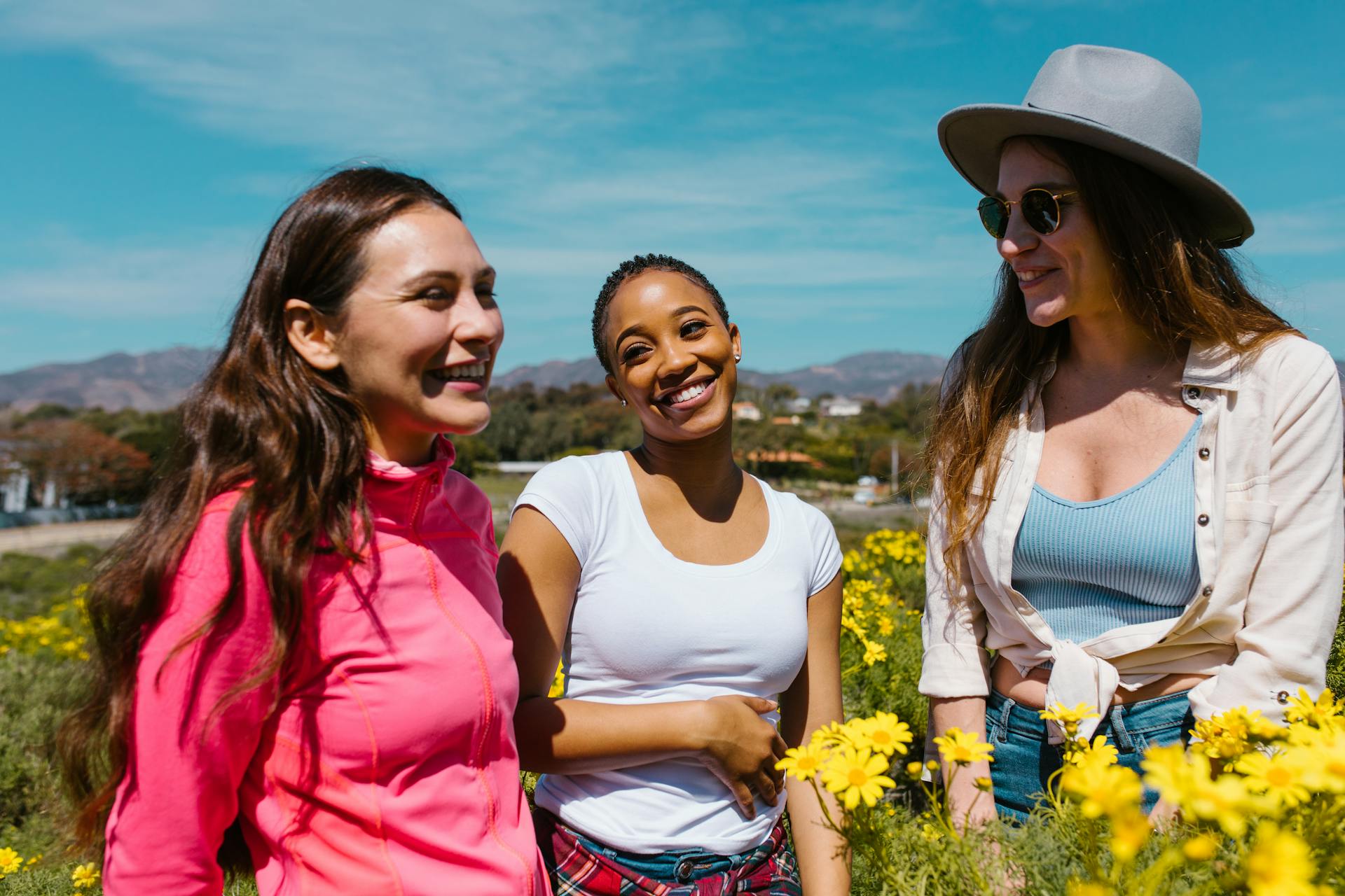 Three friends smiling and laughing together outdoors among yellow wildflowers on a sunny day.